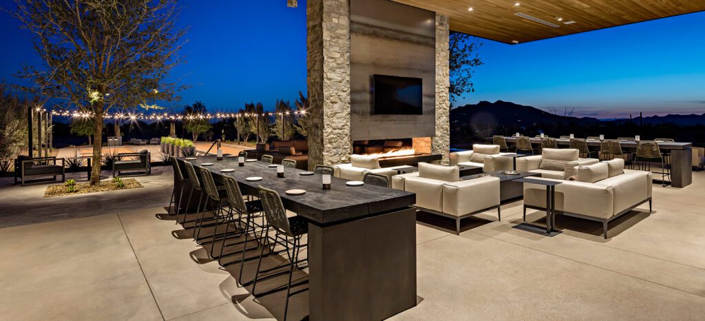 An elegant outdoor patio area at dusk, featuring a bar seating area, comfortable lounge furniture, and a fireplace with a mountainous backdrop at Seven Desert Mountain.